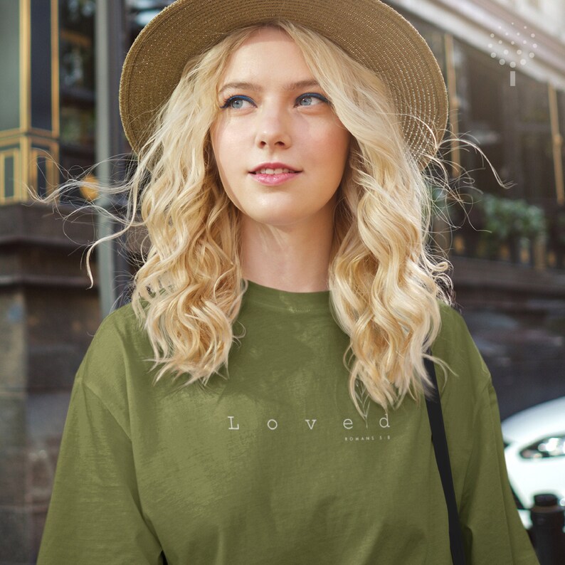 May include: A woman wearing a green long-sleeved shirt with the text "Loved Romans 5:8" printed on the front. She is wearing a straw hat and has blonde hair.