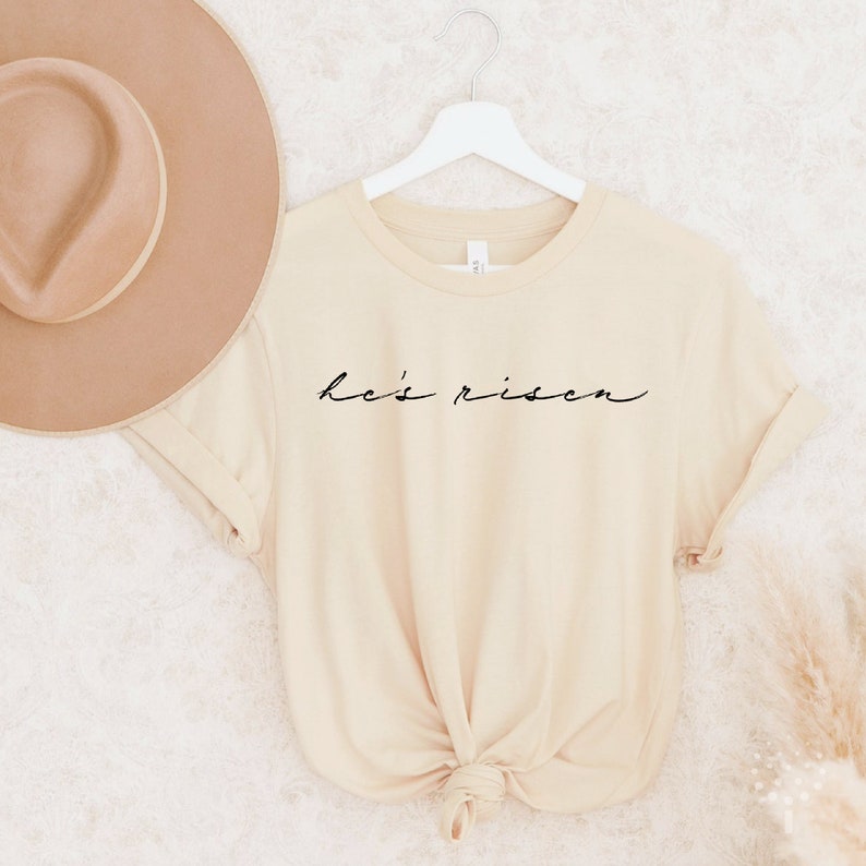 以下が含まれることがあります： 黒字で「He's Risen」と書かれたクリーム色のTシャツ。白いハンガーにかけられており、背景にはベージュの帽子があります。Tシャツはウエストで結ばれています。