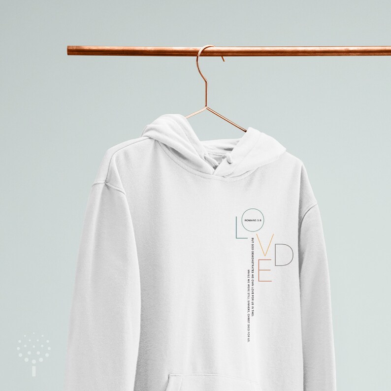 May include: White hoodie with a graphic design of the word "LOVED" in different colors and a quote from Romans 5:8. The quote reads "But God demonstrates his own love for us in this: While we were still sinners, Christ died for us."