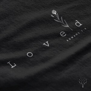 May include: Black t-shirt with white text that reads "Loved" and a small floral design. The text "Romans 5:8" is also printed in white below the floral design.