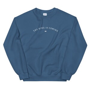 Puede incluir: Una sudadera azul con el texto "THE KING IS COMING" en letras blancas y un s&iacute;mbolo de corona.