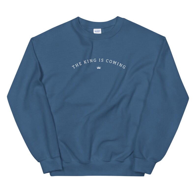 Puede incluir: Una sudadera azul con el texto "THE KING IS COMING" en letras blancas y un s&iacute;mbolo de corona.