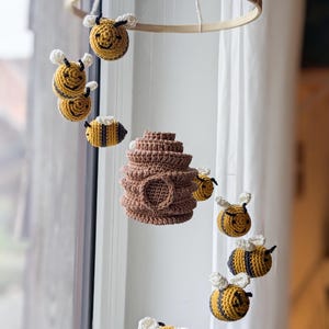 Op de afbeelding: Een gehaakte babymobiel met bijenmotief, met een bruine bijenkorf en gele en zwarte bijen die aan een houten ring hangen.