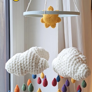 Könnte beinhalten: Ein weißer gehäkelter Baby-Mobile mit zwei Wolken, einer gelben Sonne und regenbogenfarbenen Regentropfen, die an den Wolken hängen.
