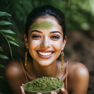 May include: A woman with green powder on her forehead smiles while holding a bowl of green powder.