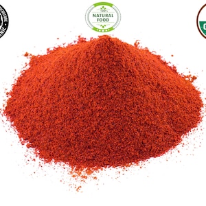Op de afbeelding: Een close-up van een hoop helderrood paprikapoeder. Het poeder is fijn gemalen en heeft een gladde textuur. Het poeder ligt op een witte achtergrond. De afbeelding bevat ook drie labels: "USDA Organic", "100% Natural Food" en "Texas Department of Agriculture Certified Organic".