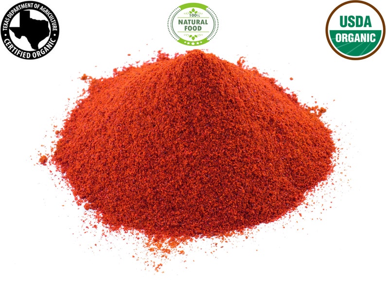 Op de afbeelding: Een close-up van een hoop helderrood paprikapoeder. Het poeder is fijn gemalen en heeft een gladde textuur. Het poeder ligt op een witte achtergrond. De afbeelding bevat ook drie labels: "USDA Organic", "100% Natural Food" en "Texas Department of Agriculture Certified Organic".