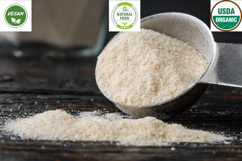 May include: A close-up of a white powder, likely tapioca starch, being poured from a metal measuring spoon onto a dark wooden surface. The powder is labeled as vegan, 100% natural food, and USDA organic.