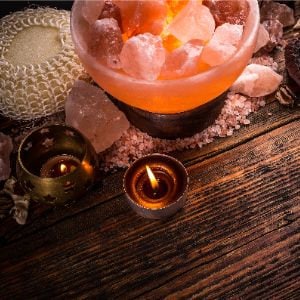 Pu&ograve; includere: Una lampada di sale dell'Himalaya con un caldo bagliore arancione, circondata da rocce di sale dell'Himalaya rosa, candele e un pouf da bagno in tessuto. La lampada di sale &egrave; in una ciotola con una base in metallo.