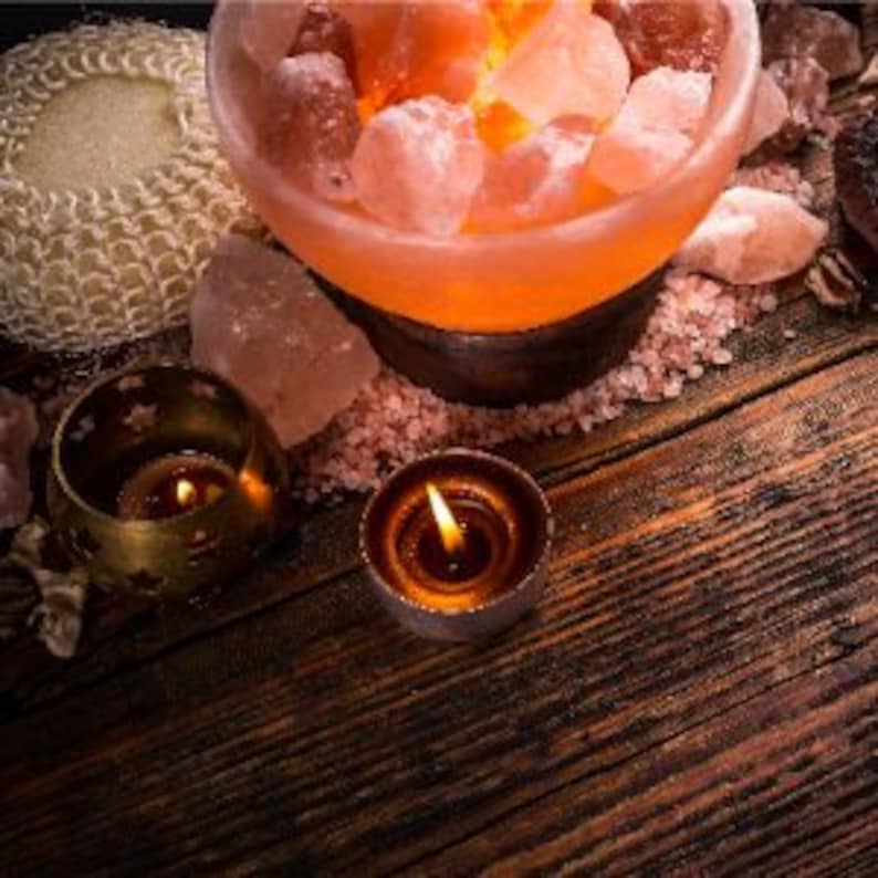 Pu&ograve; includere: Una lampada di sale dell'Himalaya con un caldo bagliore arancione, circondata da rocce di sale dell'Himalaya rosa, candele e un pouf da bagno in tessuto. La lampada di sale &egrave; in una ciotola con una base in metallo.