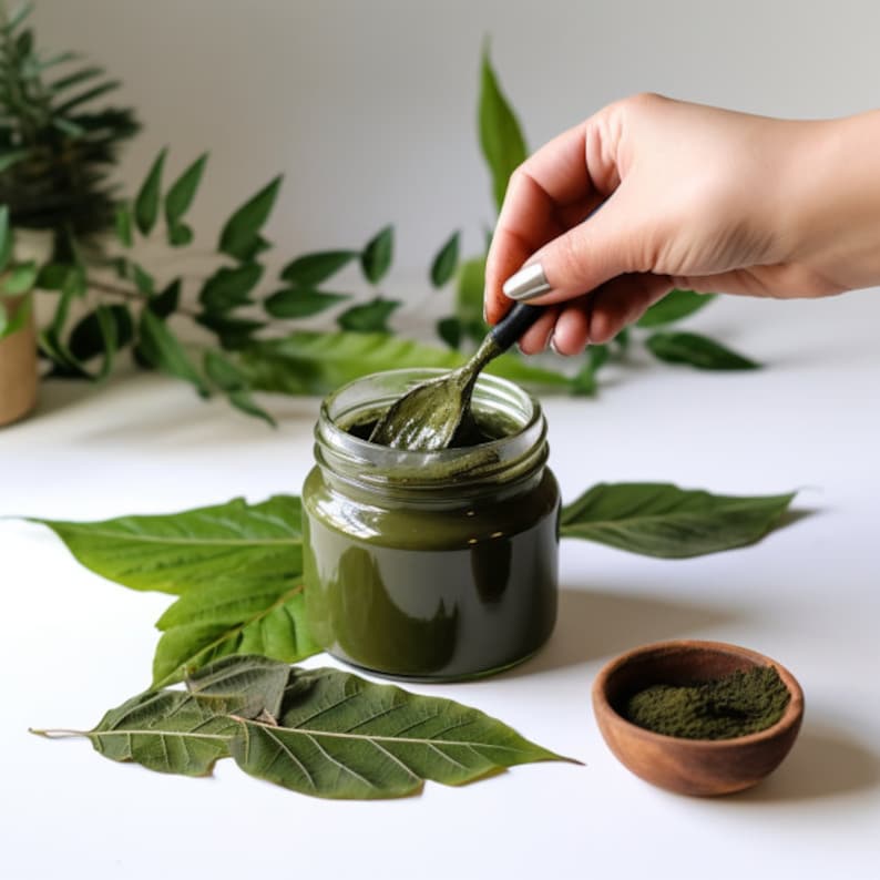 May include: A glass jar filled with a green herbal paste, with a spoon scooping out some of the paste. The jar is sitting on a white surface with green leaves scattered around it. A small wooden bowl with green powder is also visible.