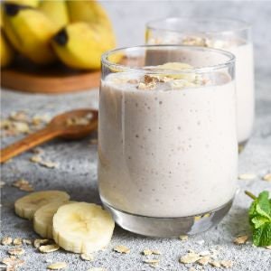 May include: Two glasses of creamy, light-coloured smoothie with a layer of oats and banana slices on top. A wooden spoon and sliced bananas are on the counter.