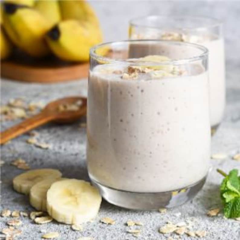 May include: Two glasses of creamy, light-coloured smoothie with a layer of oats and banana slices on top. A wooden spoon and sliced bananas are on the counter.