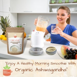 May include: A brown paper bag with a white label that reads "ORGANIC ASHWAGANDHA POWDER". The bag is sitting on a white worktop next to a blender with a glass jug filled with a light orange smoothie. A woman is holding a glass of the smoothie and smiling. The text "Enjoy A Healthy Morning Smoothie With Organic Ashwagandha" is written in brown text on a light brown background.