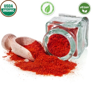 Op de afbeelding: Een glazen pot gevuld met helderrood paprikapoeder, met een houten schep en een kleine hoeveelheid gemorst poeder. De pot heeft een groen label met de tekst "Green Energy". De afbeelding bevat ook een groen USDA Organic-label en een groen Vegan-label.
