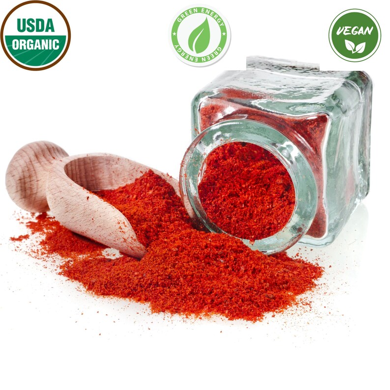 Op de afbeelding: Een glazen pot gevuld met helderrood paprikapoeder, met een houten schep en een kleine hoeveelheid gemorst poeder. De pot heeft een groen label met de tekst "Green Energy". De afbeelding bevat ook een groen USDA Organic-label en een groen Vegan-label.