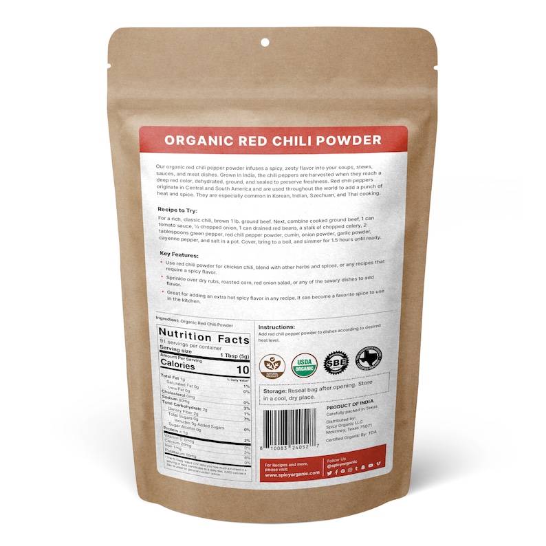 Op de afbeelding: Een bruine papieren zak met een label waarop "Organic Red Chili Powder" staat. Het label bevat een voedingssamenstelling, ingredi&euml;nten, instructies en bewaarinformatie. De zak heeft ook USDA Organic en SBE certificeringen.