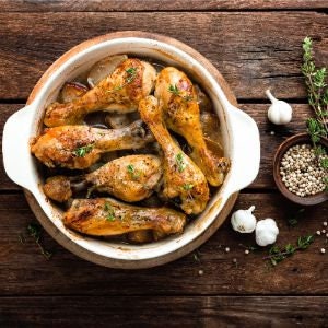 May include: A white ceramic baking dish filled with roasted chicken legs, garlic cloves, and peppercorns. The dish is on a wooden surface.
