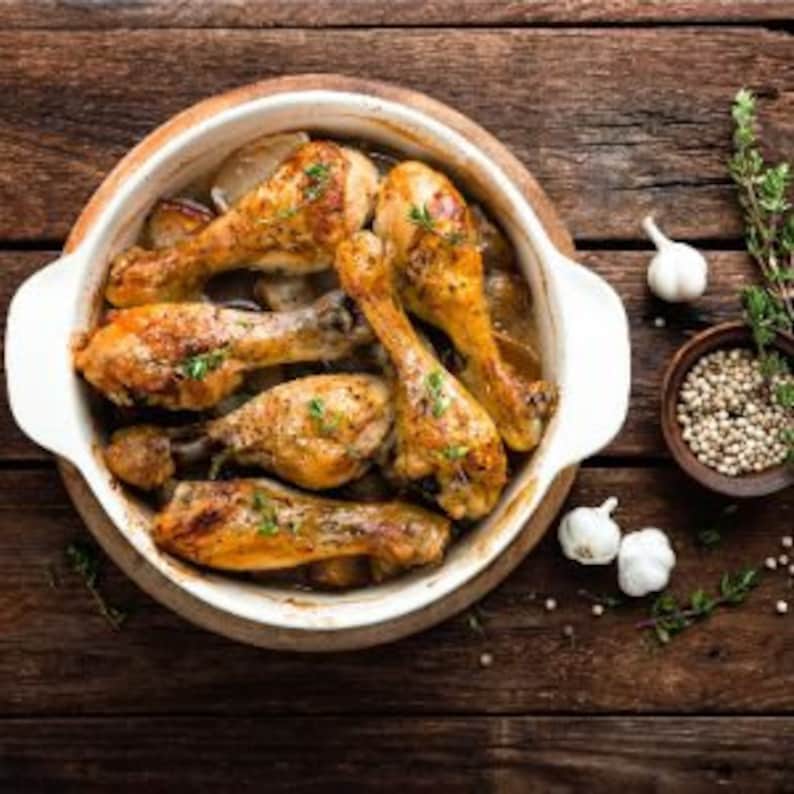 May include: A white ceramic baking dish filled with roasted chicken legs, garlic cloves, and peppercorns. The dish is on a wooden surface.