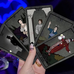 May include: A hand holds a fan of tarot cards with illustrations in shades of gray, black, and red. The cards feature figures and scenes, including a clown, a woman, and a figure in a top hat. The card labeled "THE FOOL" is visible.