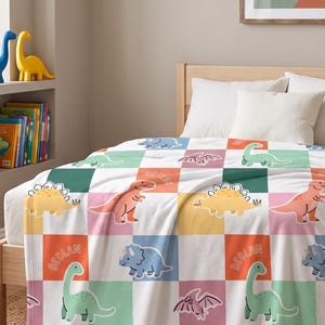 May include: A checkered throw blanket featuring a repeating pattern of various colourful dinosaurs and the name Declan printed in a playful font. The blanket displays squares in shades of green, orange, blue, and pink, covering a bed with white pillows.