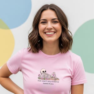 May include: A pale pink t-shirt with a skeleton graphic and the words "that wasn't very evidence based of you". The illustration shows a skeleton reclining amongst flowers. The t-shirt has a crew neck.