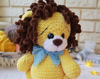 May include: A crocheted yellow lion with brown yarn hair and a blue lace bow. The lion is standing on a white surface with gold star lights.