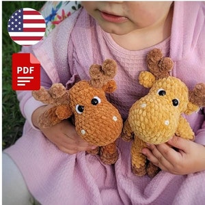 Puede incluir: Patrón PDF para tejer dos peluches de alces. Un alce es marrón y el otro es de color marrón claro. Los peluches de alces están siendo sostenidos por un niño que lleva un vestido rosa.