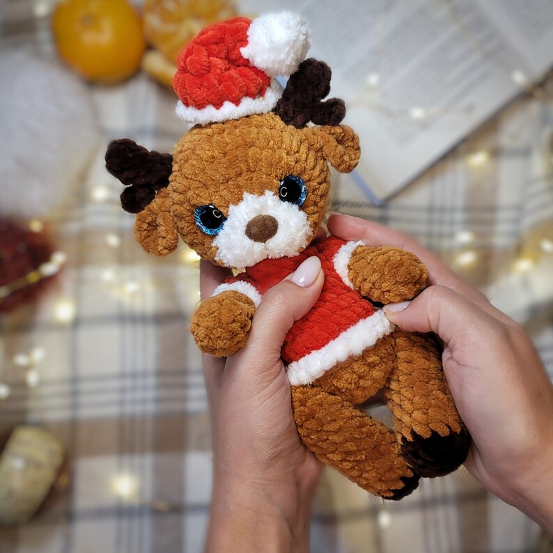 May include: A brown crocheted reindeer wearing a red and white Santa hat. The reindeer has blue eyes and a white face. The reindeer is being held by a person's hands.
