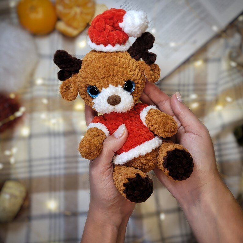 May include: A crocheted reindeer toy wearing a red and white Santa hat. The reindeer has brown fur, blue eyes, and a red and white Santa suit.