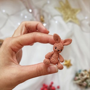 May include: A small, crocheted bunny in a warm, muted brown color. The bunny has long ears, small black eyes, and is held in a hand. The handmade amigurumi is a charming, miniature toy.
