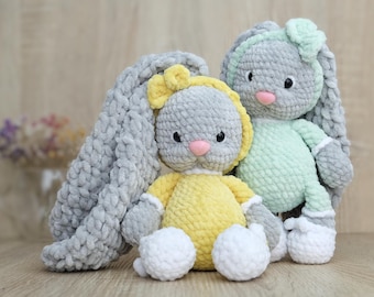 May include: Two crocheted bunny plush toys. One bunny is gray with a yellow bow and a yellow body. The other bunny is green with a gray bow. Both bunnies have white crocheted feet.
