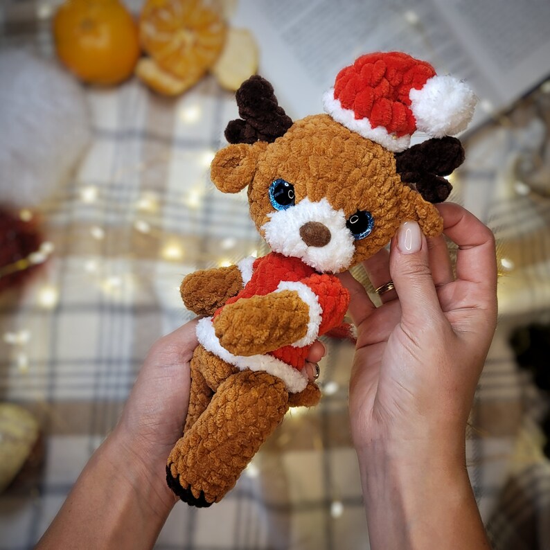 May include: A brown crocheted reindeer wearing a red and white Santa hat. The reindeer has blue eyes and a white fluffy nose.