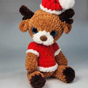 May include: A brown crocheted reindeer wearing a red and white Santa hat and a red crocheted sweater. The reindeer is sitting on a white surface.