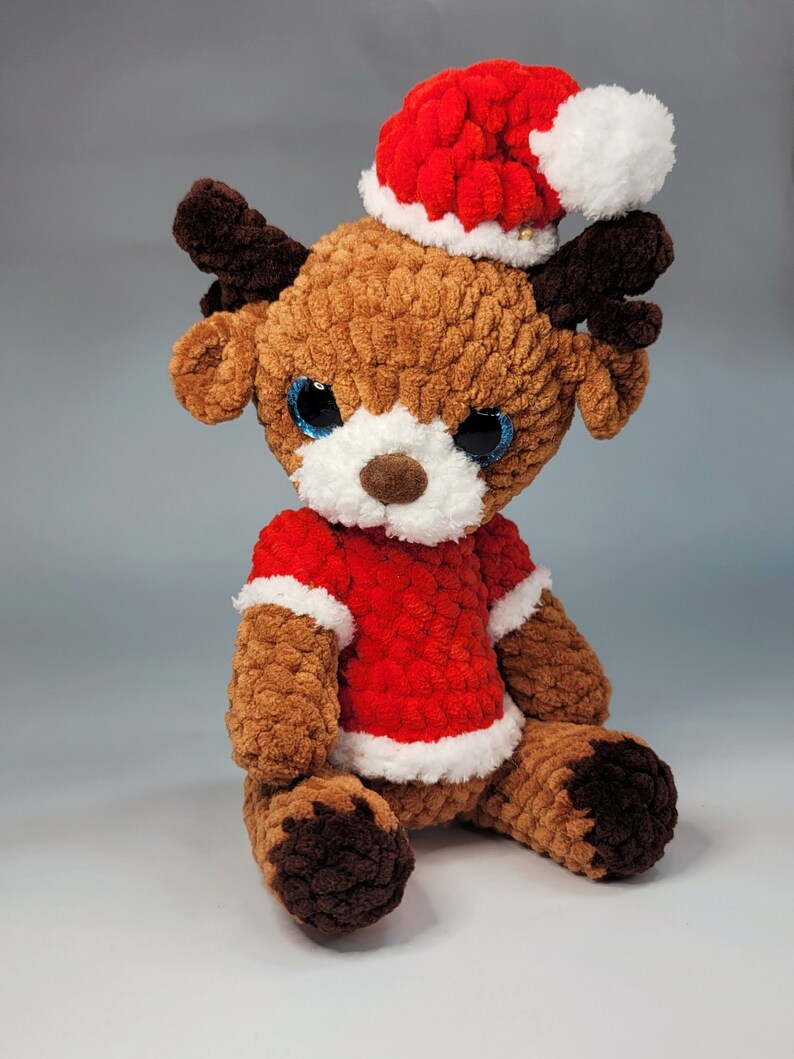 May include: A brown crocheted reindeer wearing a red and white Santa hat and a red crocheted sweater. The reindeer is sitting on a white surface.