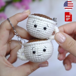 May include: Two crocheted white coffee cup keyrings, each with a smiling face and filled with brown yarn, are connected by a silver chain. The keyrings are held up by a person's hands. The coffee cups are approximately 5 cm tall.