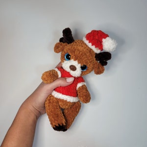 May include: A brown crocheted reindeer wearing a red and white Santa hat. The reindeer has blue eyes and a white nose.