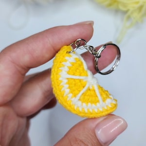 May include: A crocheted lemon slice keyring. The keyring features a bright yellow exterior with white detailing, mimicking the segments of a lemon. The keyring is attached to a silver-coloured metal ring.