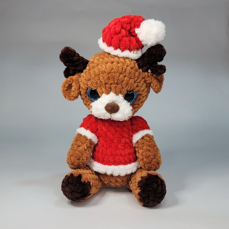May include: A brown and white crocheted reindeer wearing a red and white Santa hat. The reindeer has blue eyes and a white nose.