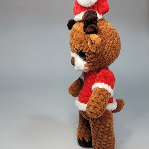 May include: A brown crocheted reindeer wearing a red and white Santa hat with a white pom-pom. The reindeer has brown antlers and black eyes.