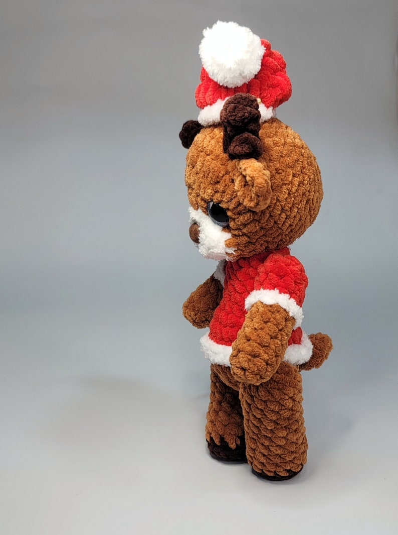 May include: A brown crocheted reindeer wearing a red and white Santa hat with a white pom-pom. The reindeer has brown antlers and black eyes.