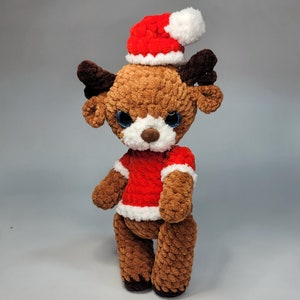 May include: A brown crocheted reindeer wearing a red and white Santa hat. The reindeer has blue eyes and a white nose.