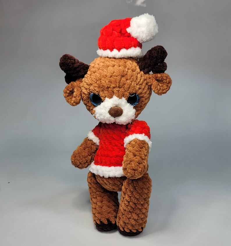 May include: A brown crocheted reindeer wearing a red and white Santa hat. The reindeer has blue eyes and a white nose.