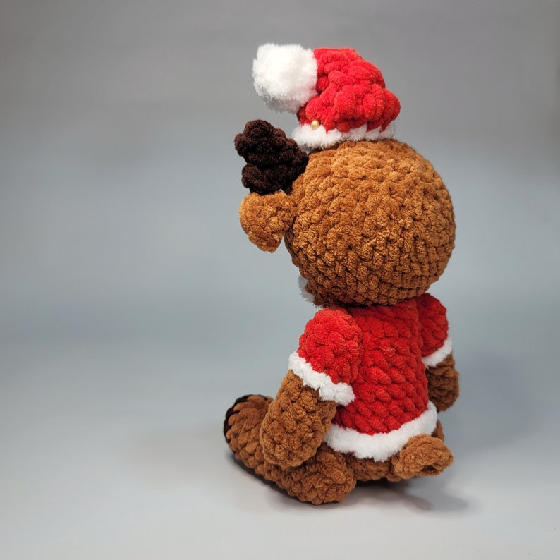 May include: A brown crocheted reindeer wearing a red and white Santa hat. The reindeer has a white trim around the hat and a white trim around the red suit.