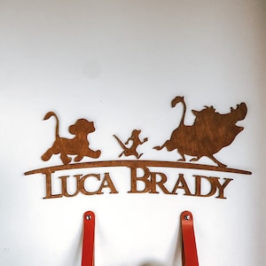 May include: Wooden wall decoration featuring the characters Simba, Timon, and Pumbaa from the Disney film The Lion King. The characters are standing on a curved wooden base with the name "Luca Brady" written below.