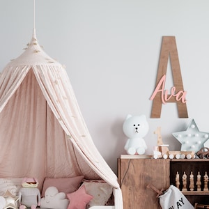 K&ouml;nnte beinhalten: Ein rosafarbenes Holz-Namensschild mit dem Namen "Ava" auf einem hellbraunen Holzgrund. Das Schild h&auml;ngt an einer wei&szlig;en Wand &uuml;ber einer Kommode aus Holz mit einem wei&szlig;en Stofftier und einem sternf&ouml;rmigen Licht.