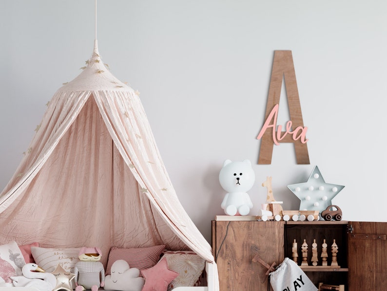 K&ouml;nnte beinhalten: Ein rosafarbenes Holz-Namensschild mit dem Namen "Ava" auf einem hellbraunen Holzgrund. Das Schild h&auml;ngt an einer wei&szlig;en Wand &uuml;ber einer Kommode aus Holz mit einem wei&szlig;en Stofftier und einem sternf&ouml;rmigen Licht.