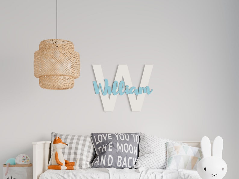 K&ouml;nnte beinhalten: Wei&szlig;er Holzbuchstabe W mit dem Namen "William" in hellblauer Farbe. Der Buchstabe h&auml;ngt an einer wei&szlig;en Wand &uuml;ber einem wei&szlig;en Bett mit Kissen und einem Stofffuchs. Eine H&auml;ngeleuchte aus Weidengeflecht h&auml;ngt &uuml;ber dem Bett.