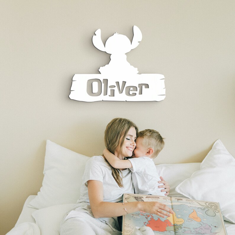 May include: White wooden sign with a cartoon Stitch character and the name "Oliver" cut out. A mother and son are sitting on a bed behind the sign.