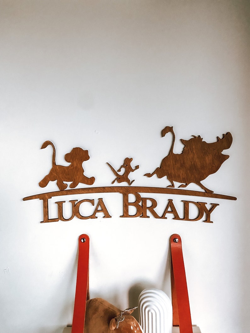 May include: Wooden wall decoration featuring the characters Simba, Timon, and Pumbaa from the Disney film The Lion King. The characters are standing on a curved wooden base with the name "Luca Brady" written below.